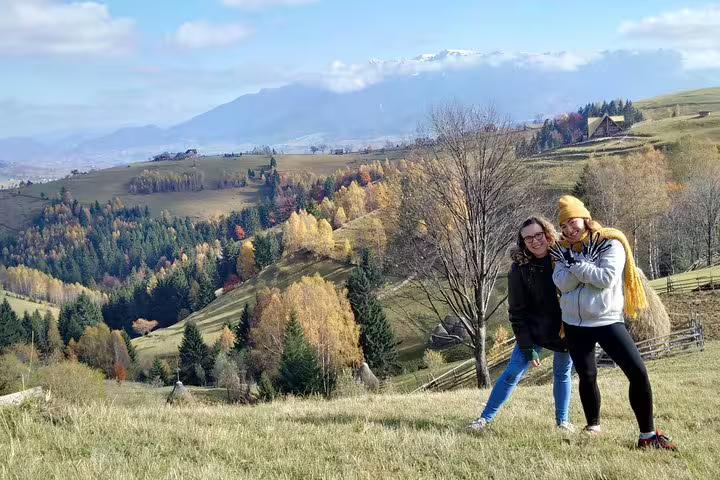 Two women posing on a hillside with colorful autumn mountain villages and Carpathian peaks near Brasov, Romania