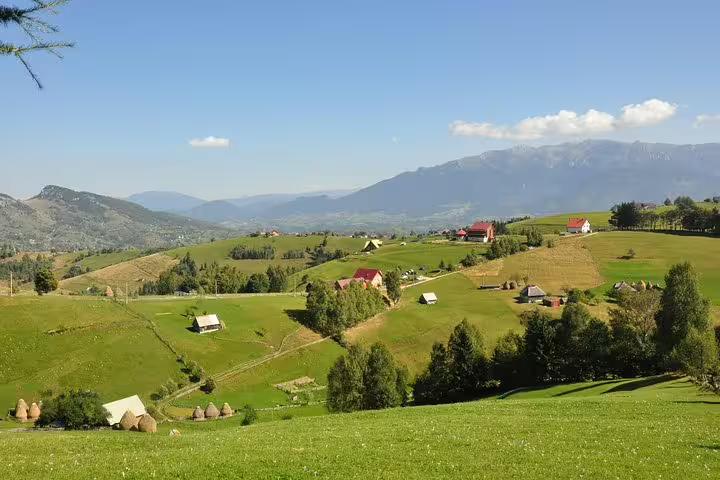 Green rolling hills with traditional Romanian farmhouses and haystacks, mountain backdrop near Brasov