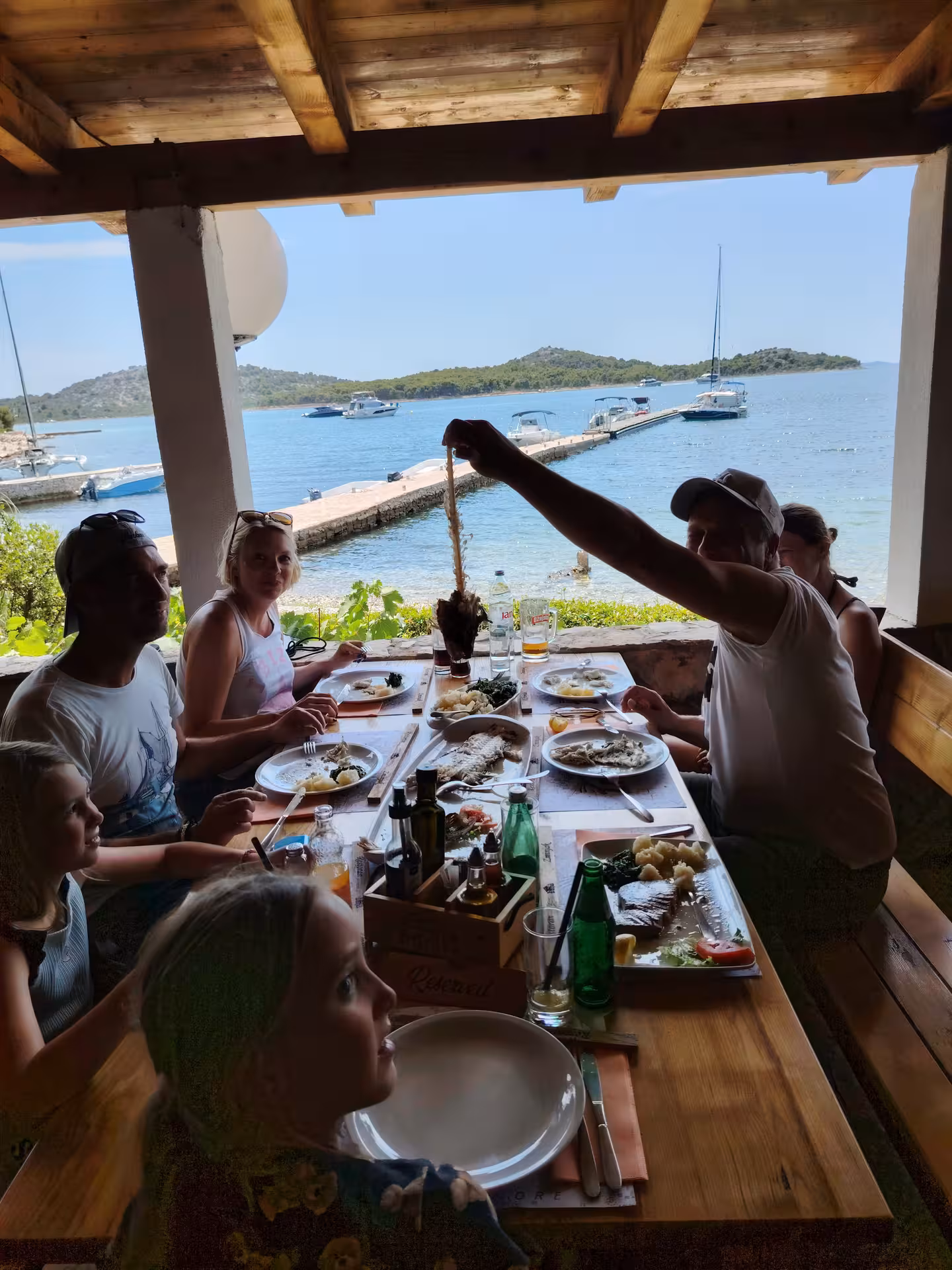 Group enjoying seaside lunch at waterfront tavern stop on sailing adventure, marina pier and anchored yachts beyond