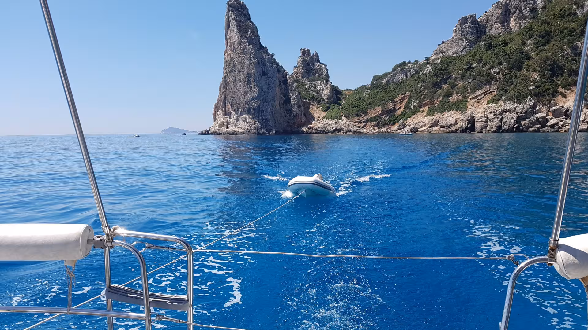 Sailing near dramatic rock formations in the Gulf of Orosei with a small dinghy in tow, under a clear blue sky.