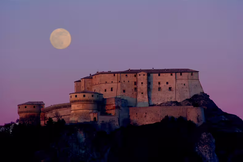 San Leo Fortress at dusk with a full moon, showcasing its majestic silhouette against a purple sky.