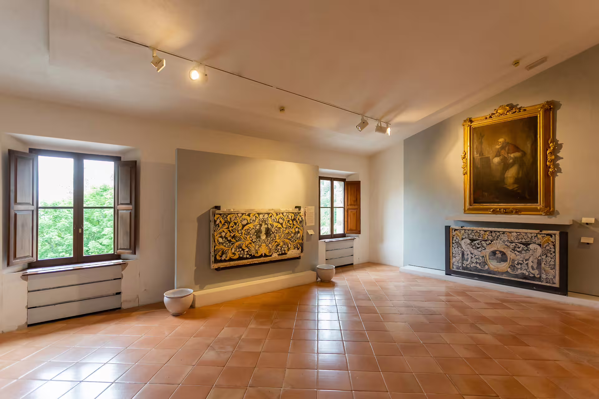 Gallery room in San Leo Fortress featuring ornate art pieces and windows overlooking scenic landscapes.