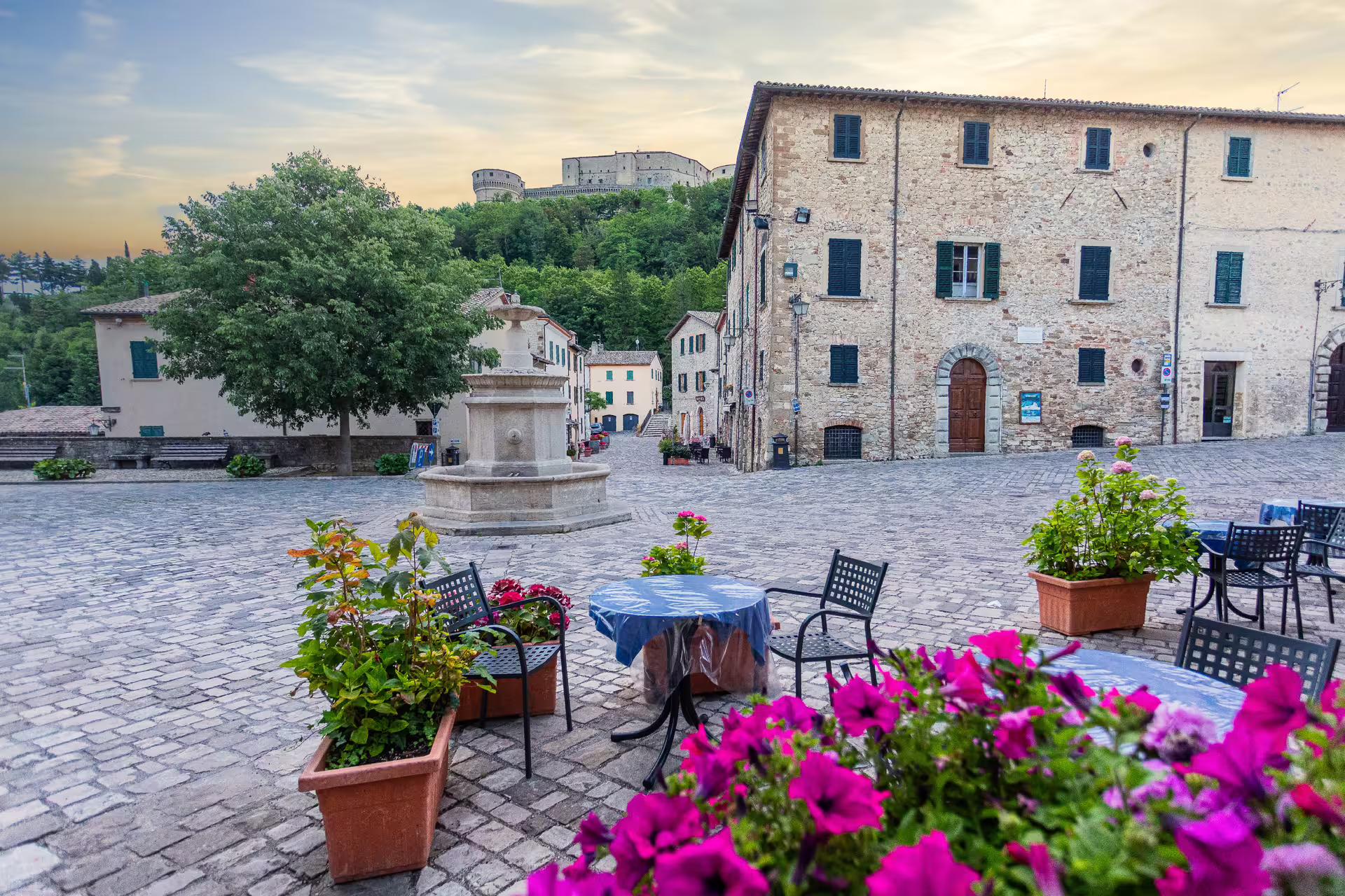 Charming San Leo square with outdoor café, vibrant flowers, and distant fortress view under a soft evening sky.