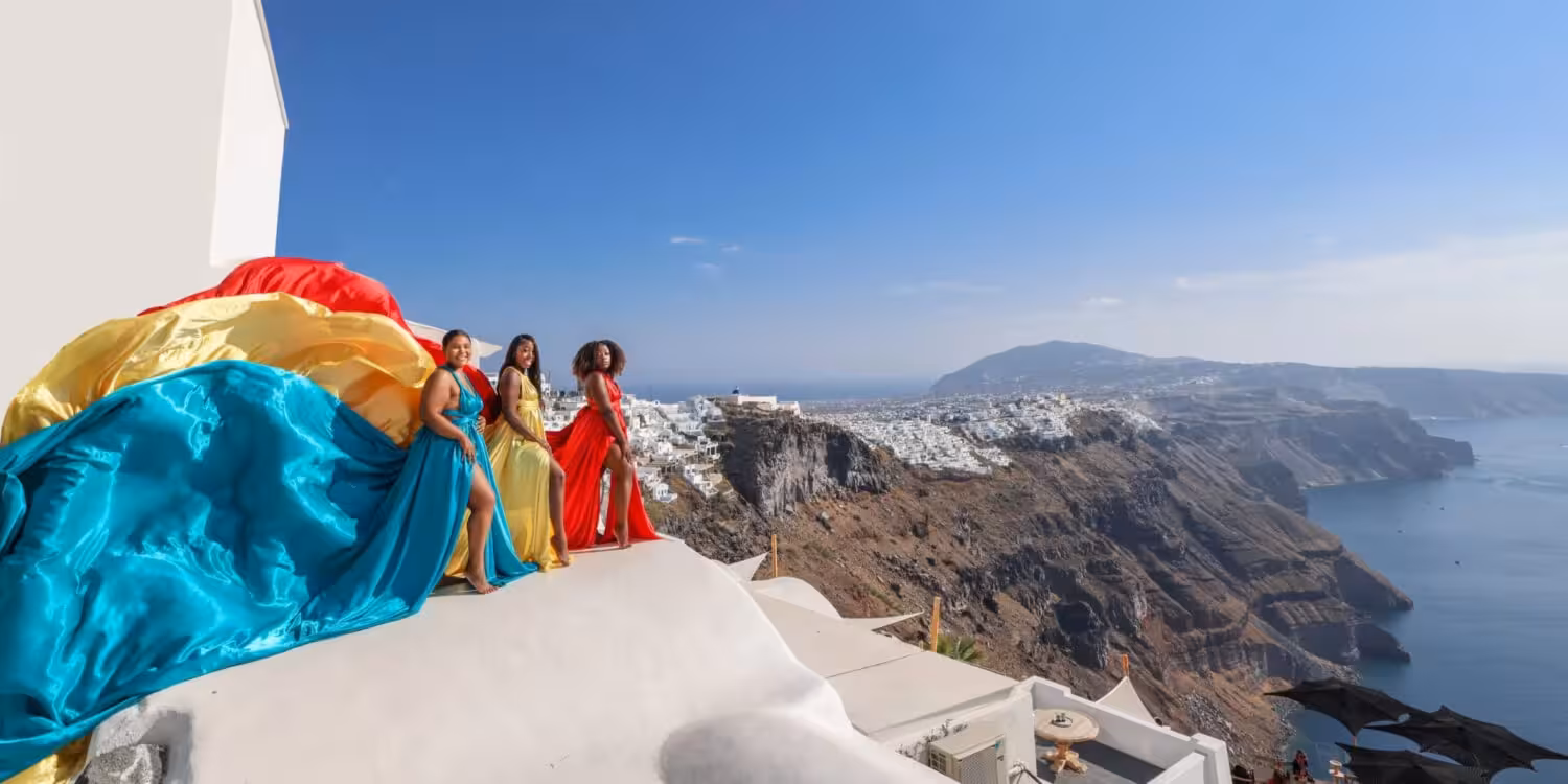 Group Santorini flying dress photoshoot on caldera cliff, colorful gowns flowing over Oia rooftops and sea
