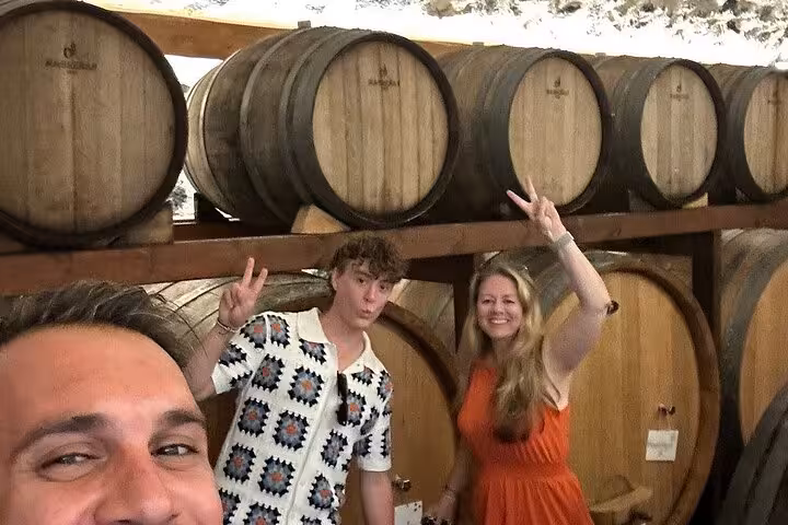 Guests posing by oak wine barrels at a Santorini winery on a private wine tasting tour with cellar visit