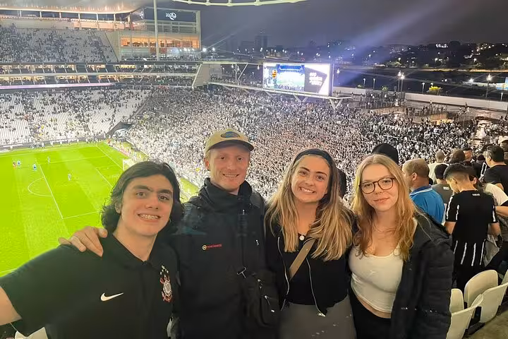 Travelers with local host at a São Paulo football match, stadium crowd behind, join a soccer game experience