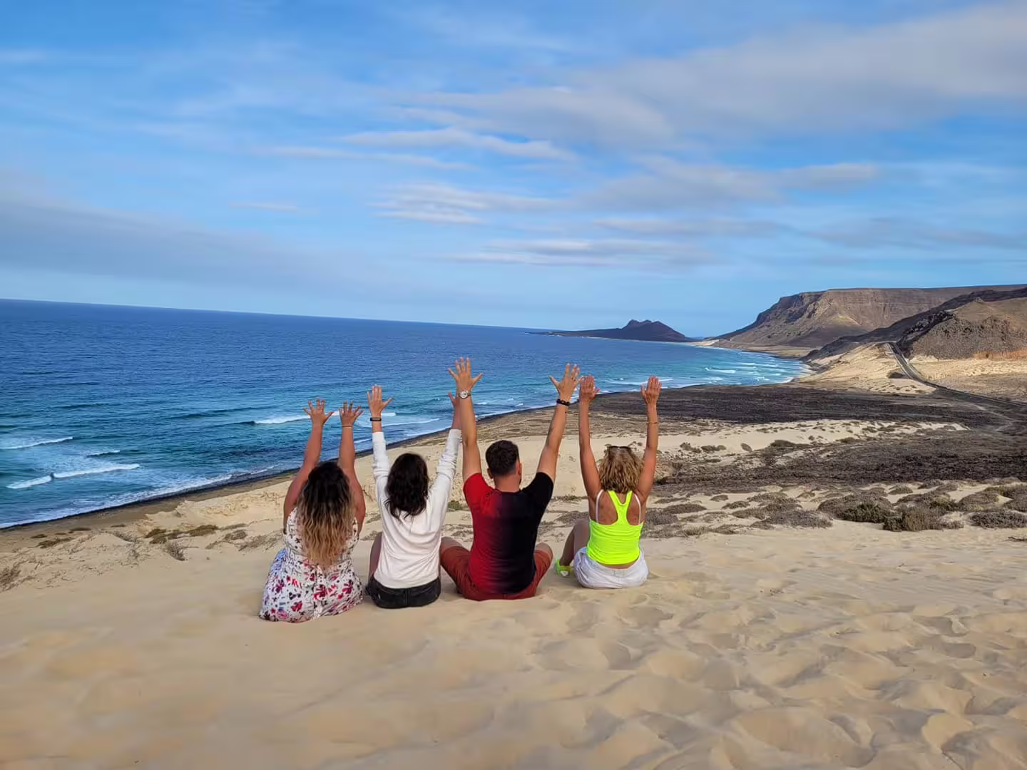Tourists enjoy stunning São Vicente beach views, sitting on sand dunes with raised arms, embracing Cape Verde's scenic beauty.