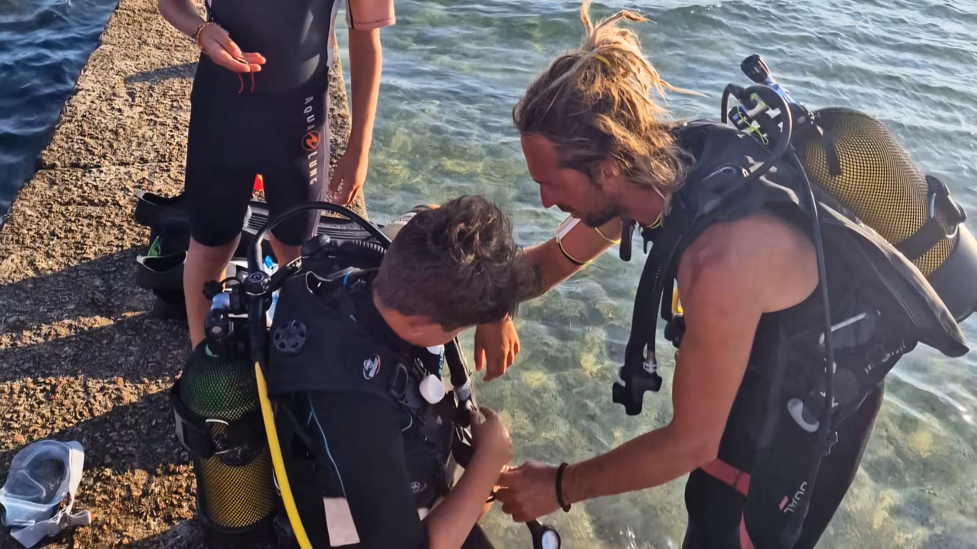 Instructor assisting a beginner diver with scuba gear by the clear waters of Argentiera, Sassari for an underwater baptism.