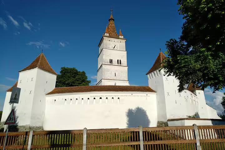 White fortified Saxon church with clock tower and corner bastions under blue sky in Transylvania