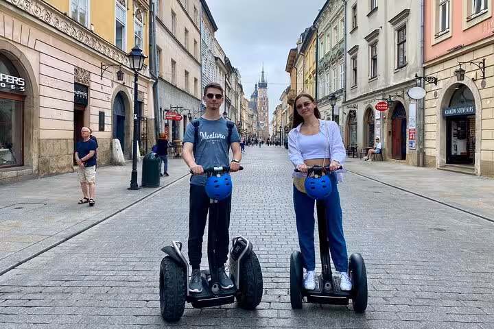 Two tourists on Segways in Krakow's vibrant Jewish Quarter, with historic buildings and a church spire in the background.