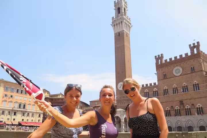 Three tourists with a flag pose in front of Siena's iconic Torre del Mangia under a clear blue sky.