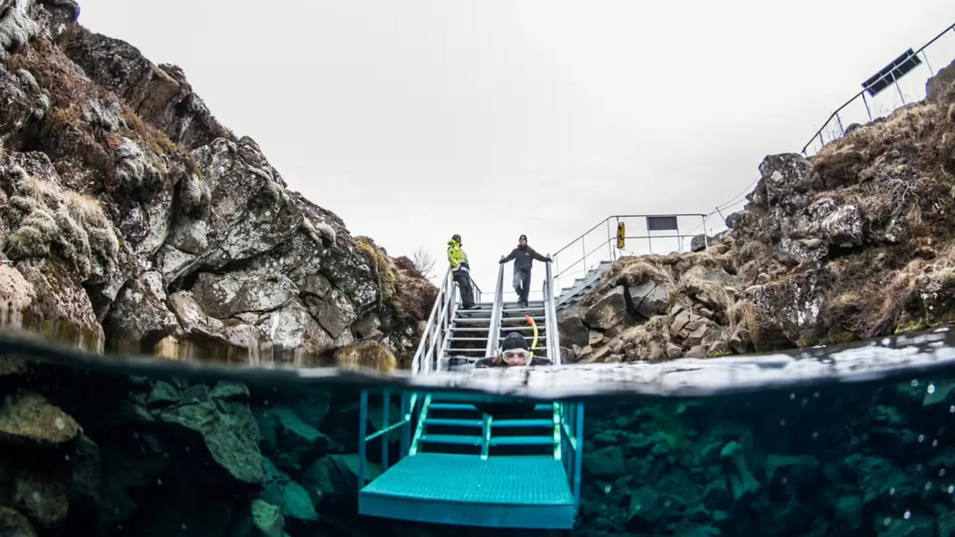 Snorkeler entering crystal clear Silfra fissure water via metal stairs with rocky canyon walls in Iceland