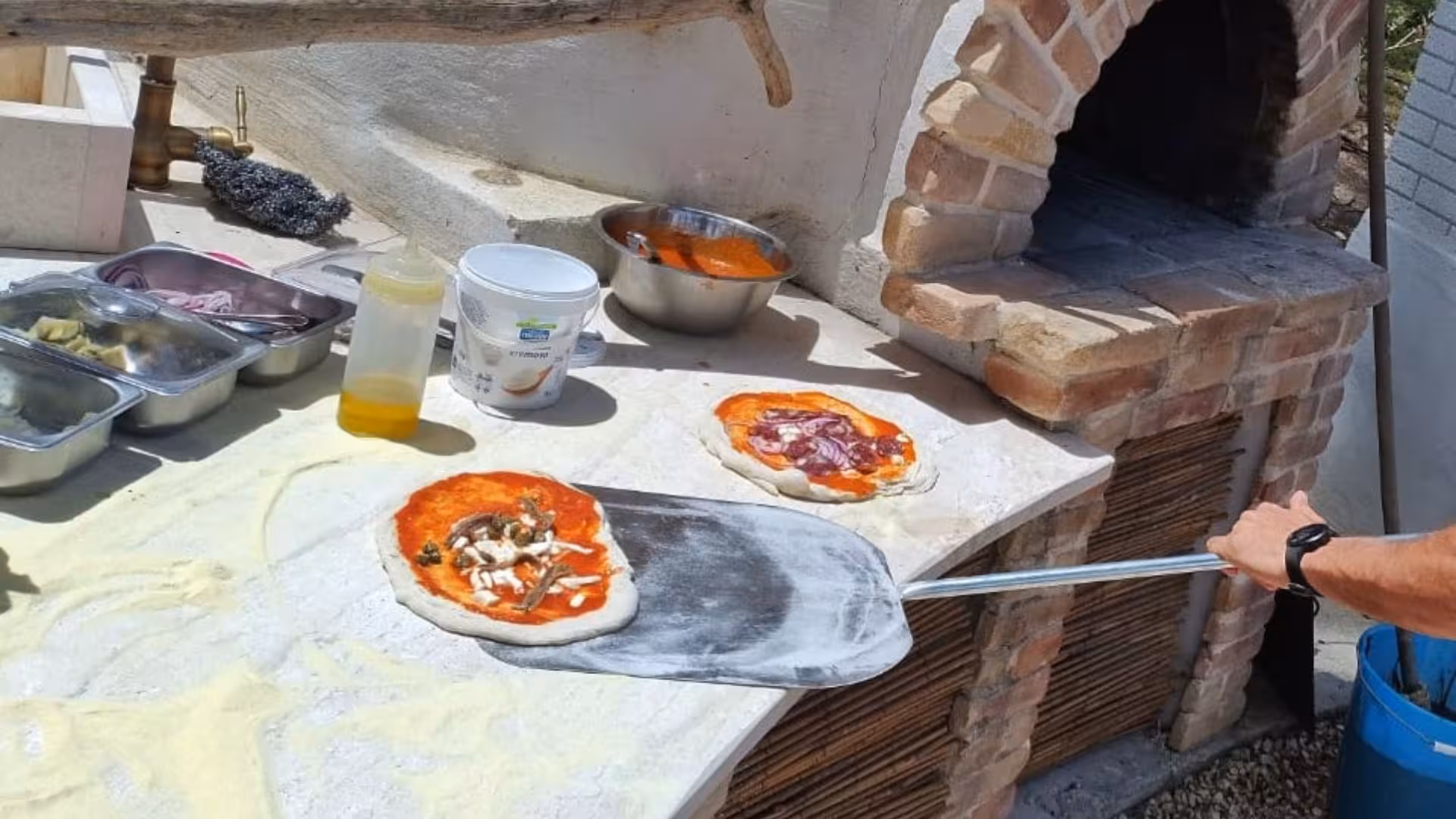 Preparing pizzas with fresh ingredients next to a rustic wood-fired oven at Sorso villa pizza workshop.