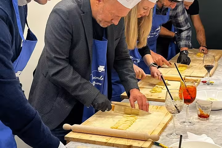 Group of people in aprons and hats cutting fresh pasta during a hands-on cooking class, with cocktails nearby.