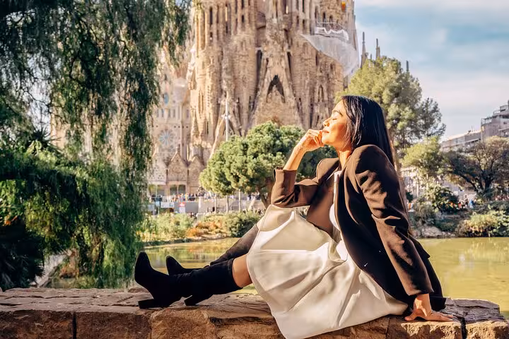 Stylish traveler posing by the pond in Plaça de Gaudí with Sagrada Família backdrop, golden-hour Barcelona photoshoot.