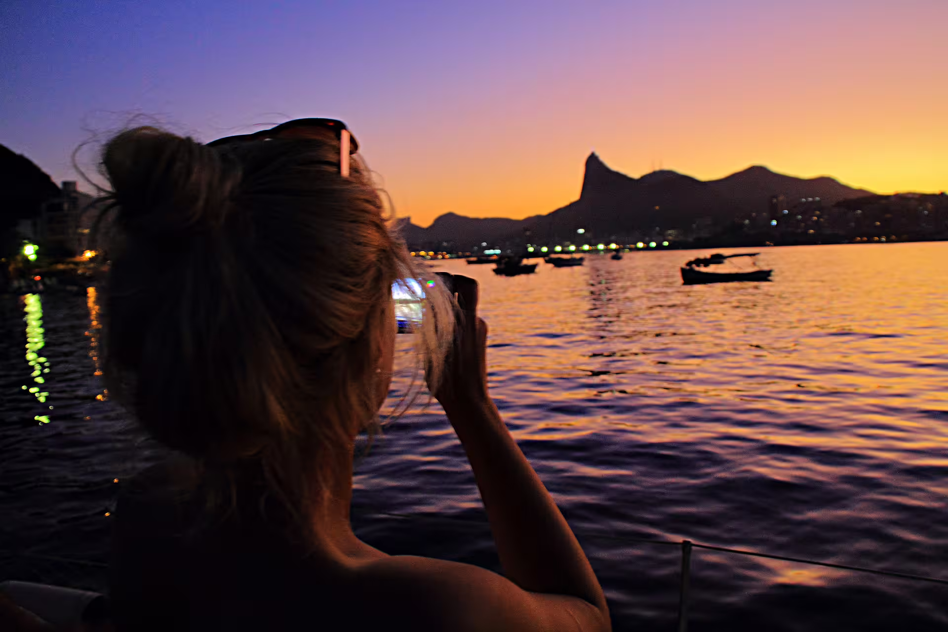 Woman capturing vibrant Rio sunset from boat with city skyline and Sugarloaf Mountain in the background.