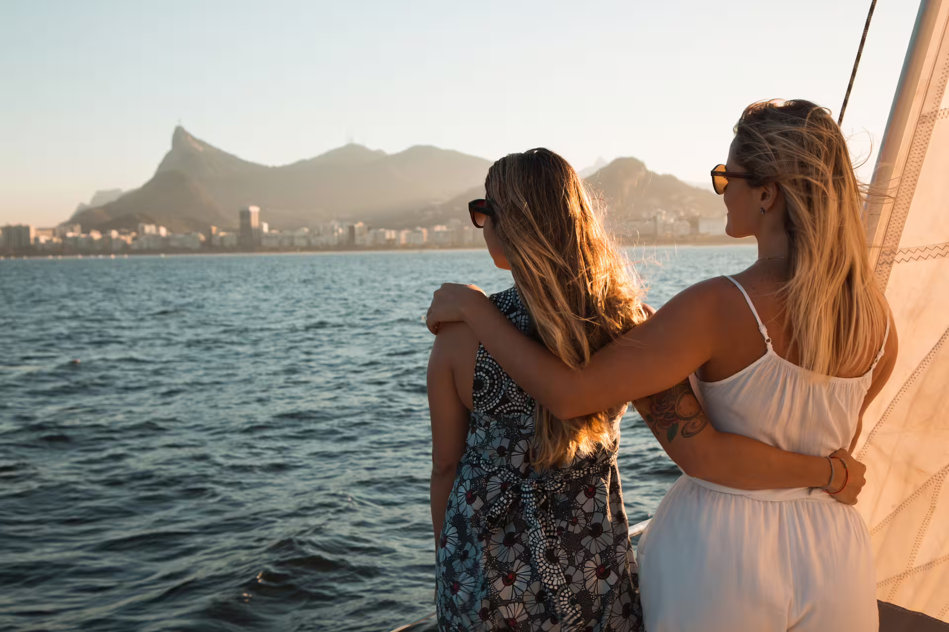 Two women embrace while admiring the scenic Rio de Janeiro skyline from a sailboat during sunset.