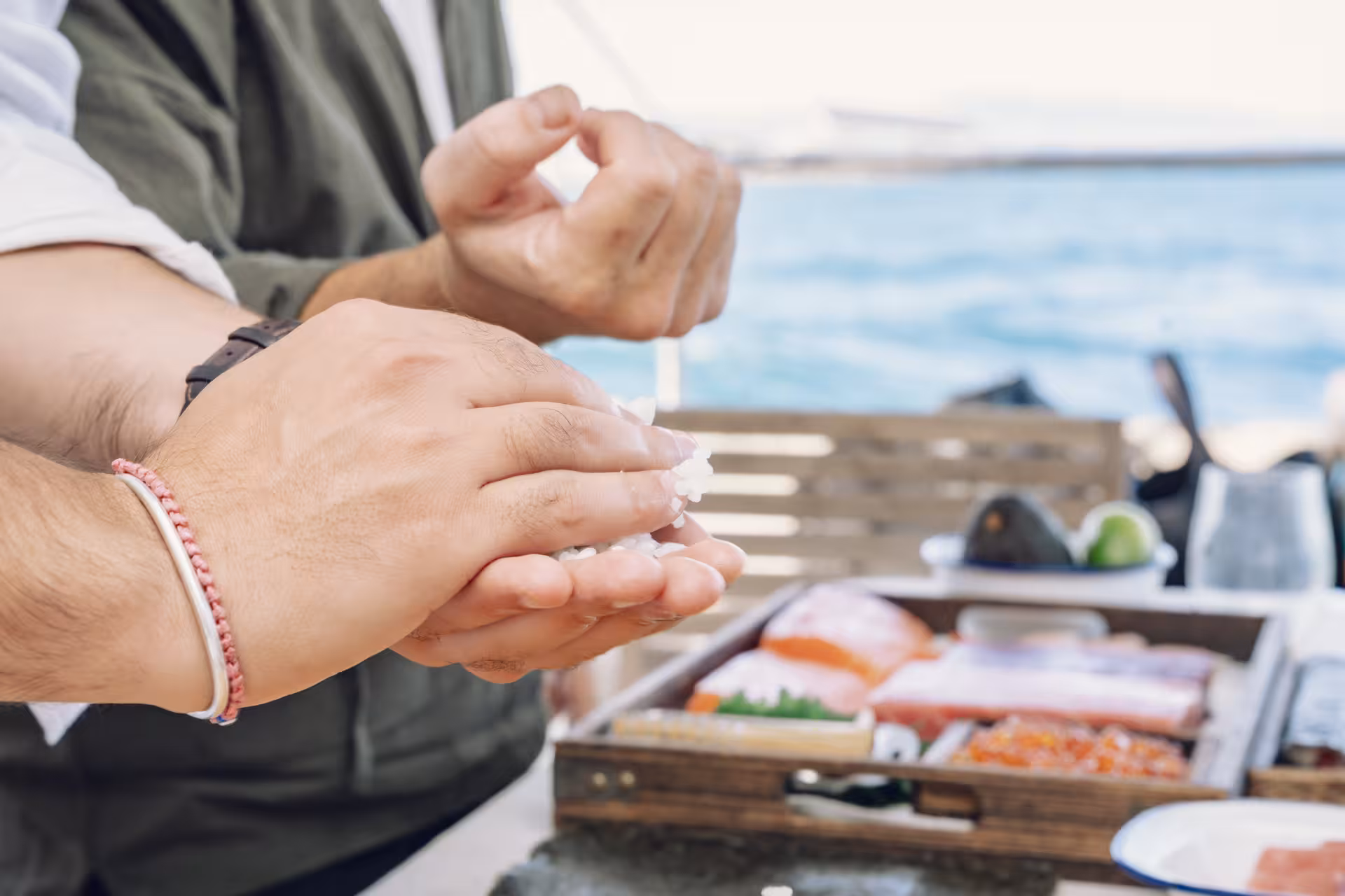 Close-up of hands shaping sushi rice in a Sushi Masterclass, with fresh fish and ingredients set up seaside