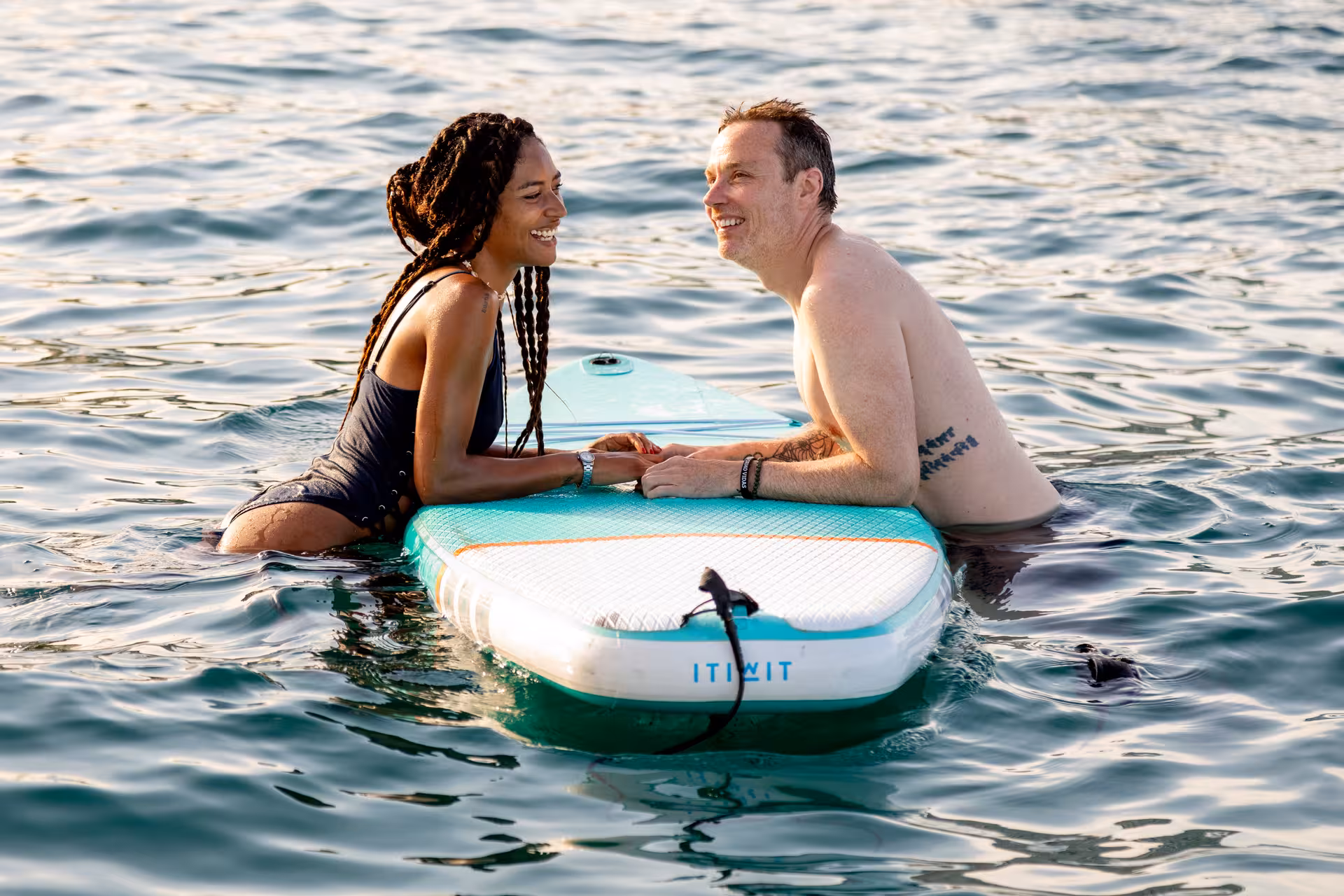 Two people smiling while resting on a paddleboard in the ocean during a sunset stand-up paddleboarding session