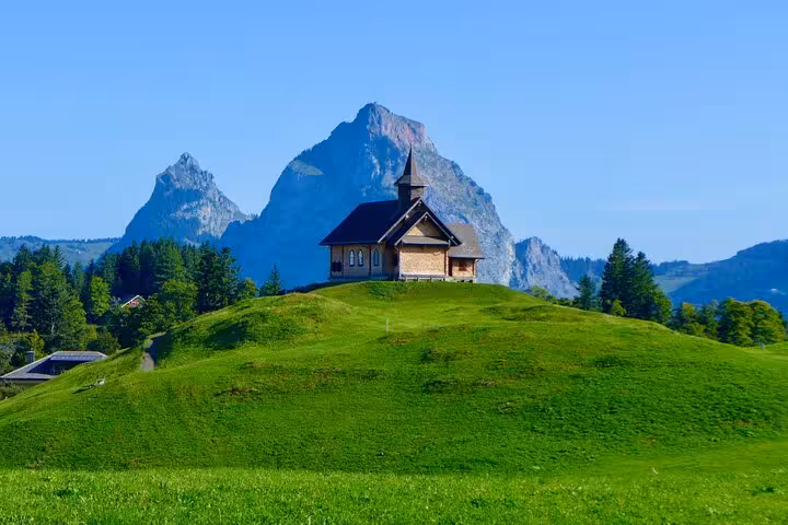 Charming chapel on a lush hill with majestic Swiss mountains in the background under a clear blue sky.