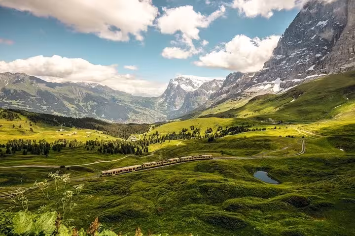 Panoramic Swiss landscape with a train traversing verdant valleys, surrounded by towering snow-capped mountains and clear skies.