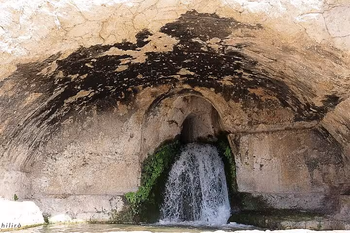 Small waterfall emerging from a stone arch in Syracuse, surrounded by lush greenery and rustic rock formations.