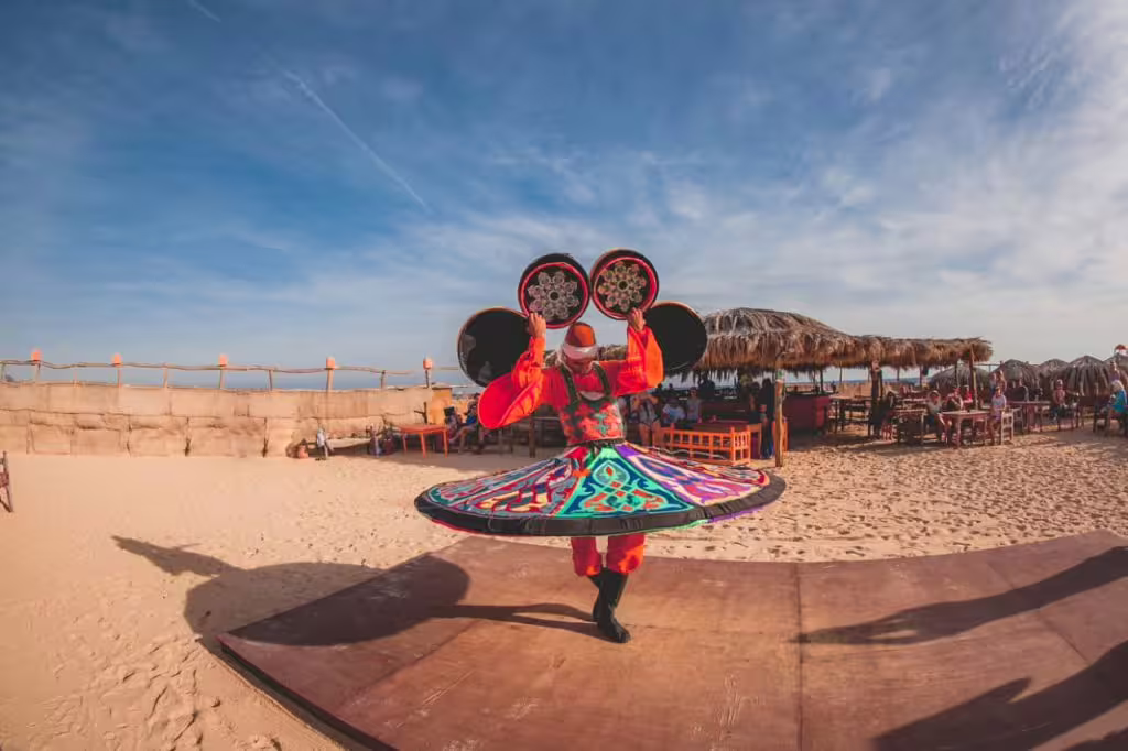 Colorful tanoura dancer performing on Paradise Island beach during day tour, with seaside huts and entertainment