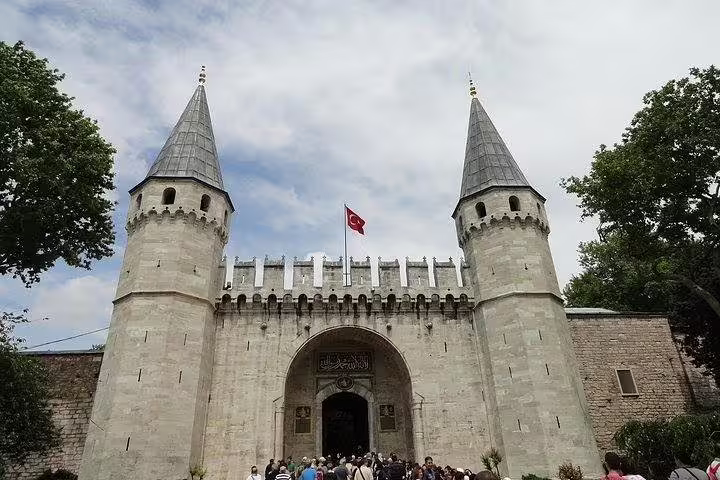 Entrance gate of Topkapi Palace in Istanbul with traditional towers and Turkish flag under a bright blue sky.
