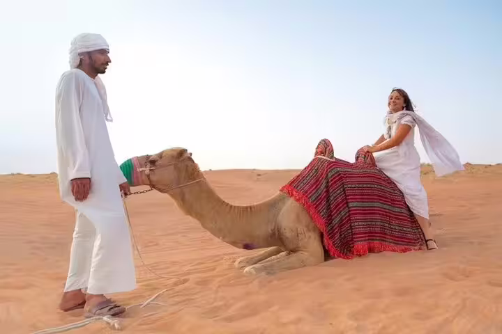 Smiling tourist enjoys a guided camel ride with local driver in traditional dress on golden dunes during Dubai desert safari