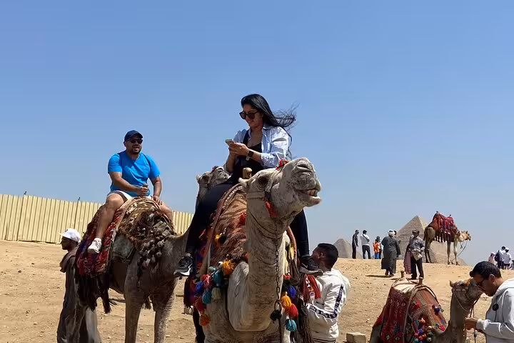 Tourists riding camels near the Giza Pyramids under a clear sky, part of a VIP Giza Pyramids and GEM day tour