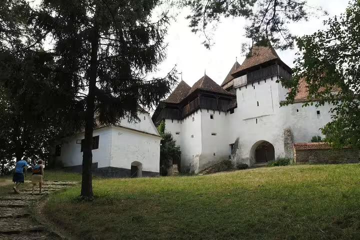 White fortified church with cone towers and wooden gallery surrounded by trees in Transylvania