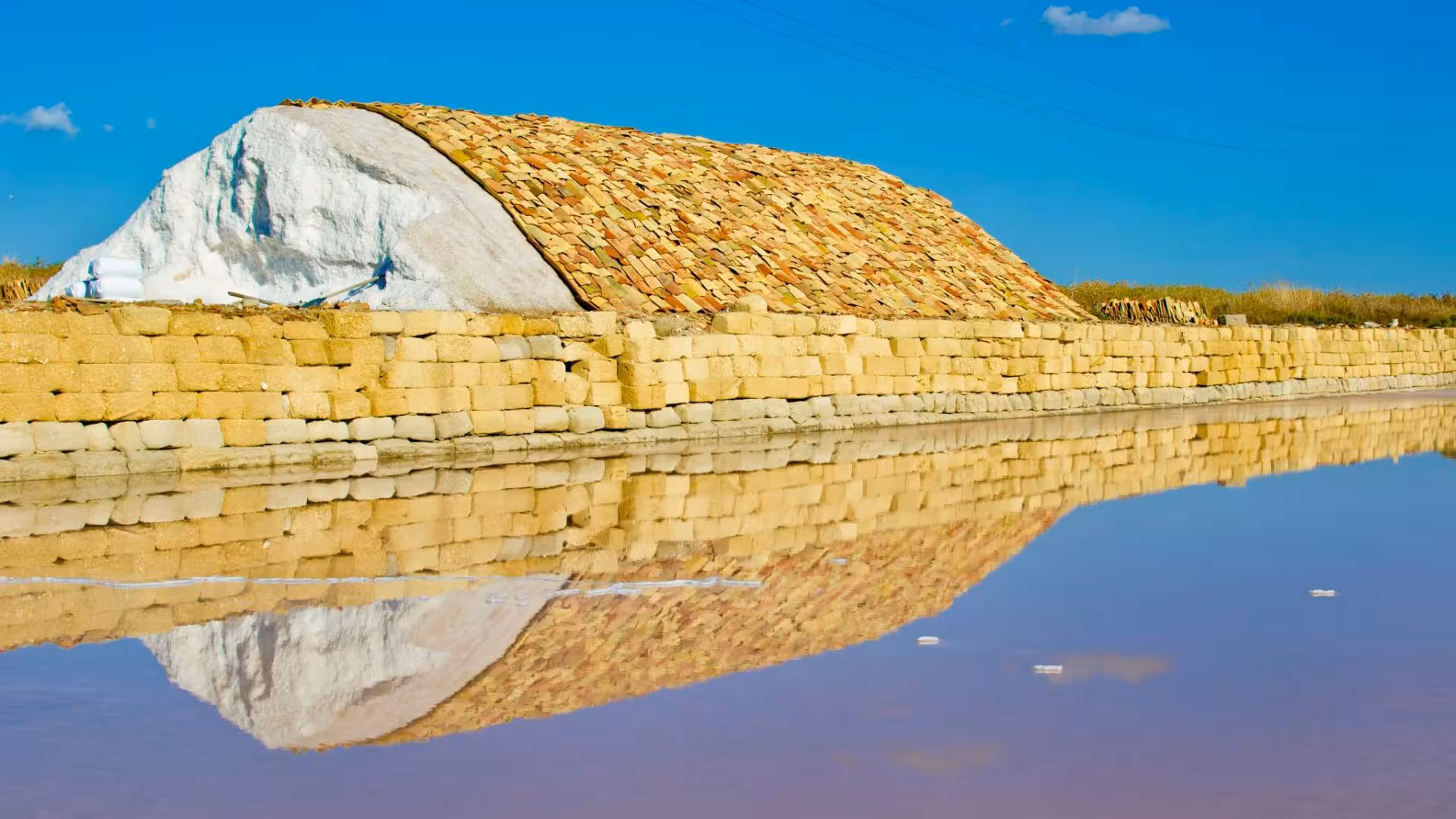 A large mound of salt covered with tiles reflects in the water at the Marsala salt pans under a clear blue sky.