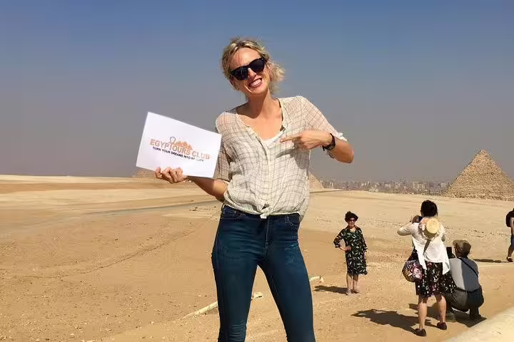 Smiling traveler holding Egypt Tours Club sign near Giza Plateau, private Pyramids day tour with Grand Egyptian Museum