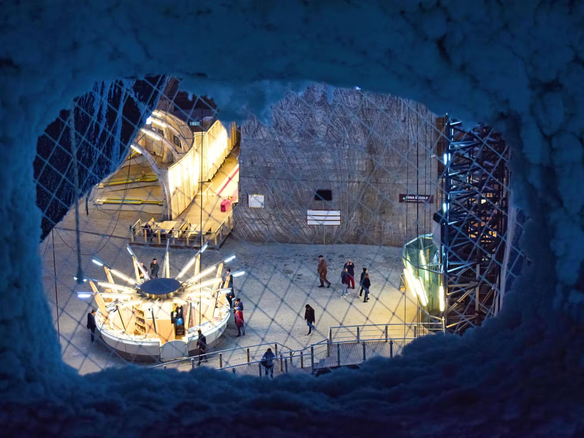 Aerial view through a salt cave opening at Turda Salt Mine showing illuminated underground attractions and visitors exploring the historic Rudolf mine chamber during a guided tour from Cluj