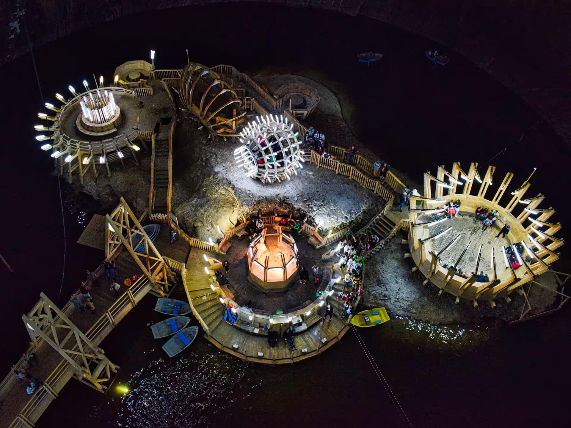 Aerial view of illuminated Turda Salt Mine interior with wooden structures, boats, and visitors exploring underground lake.