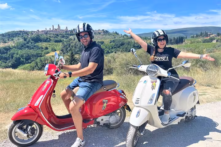 A couple riding Vespas with a panoramic view of the Tuscan countryside on a sunny day.