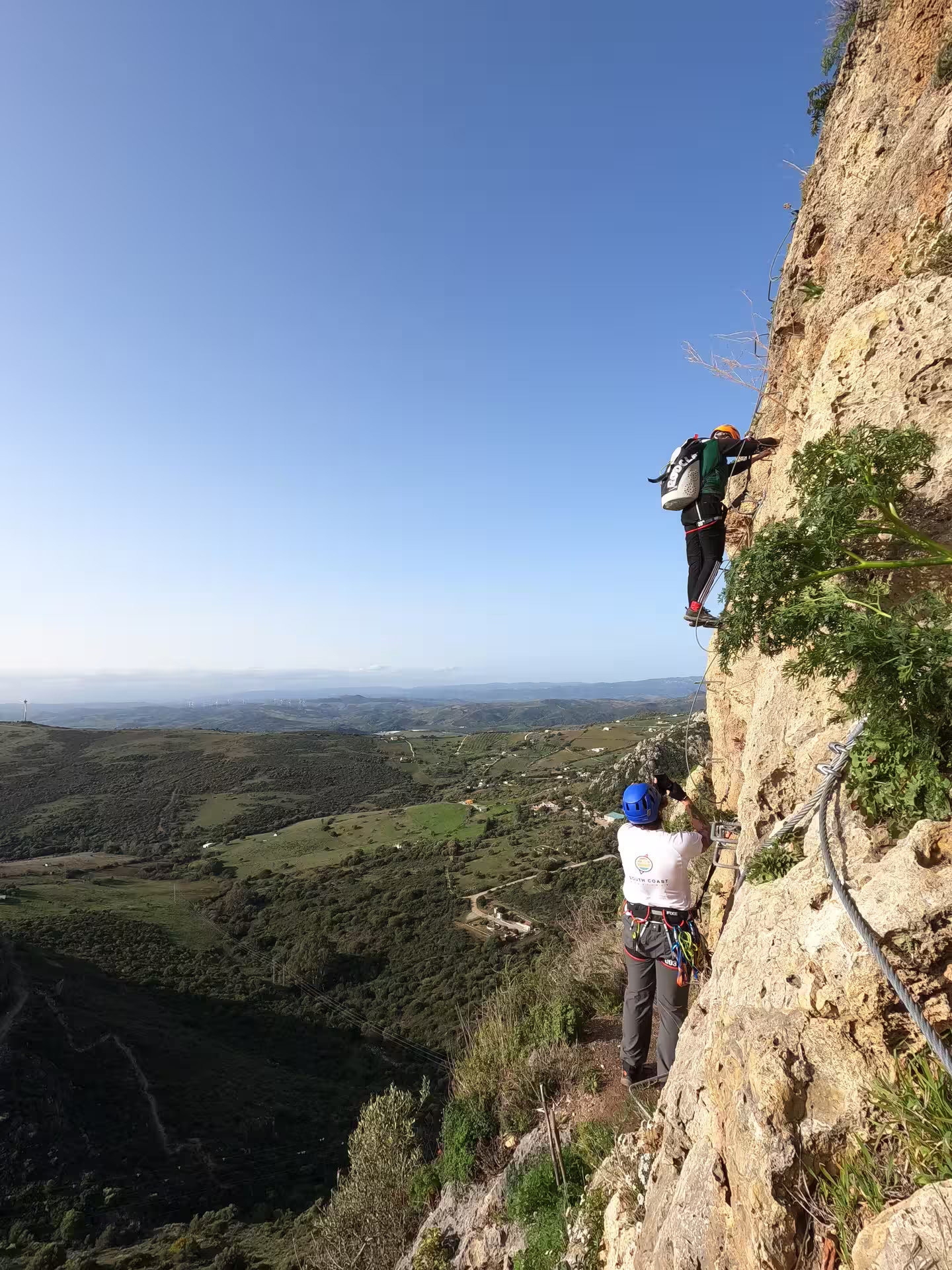 Thrill-seekers climbing a steep Via Ferrata in Casares, Spain, with breathtaking panoramic views of lush landscapes below.