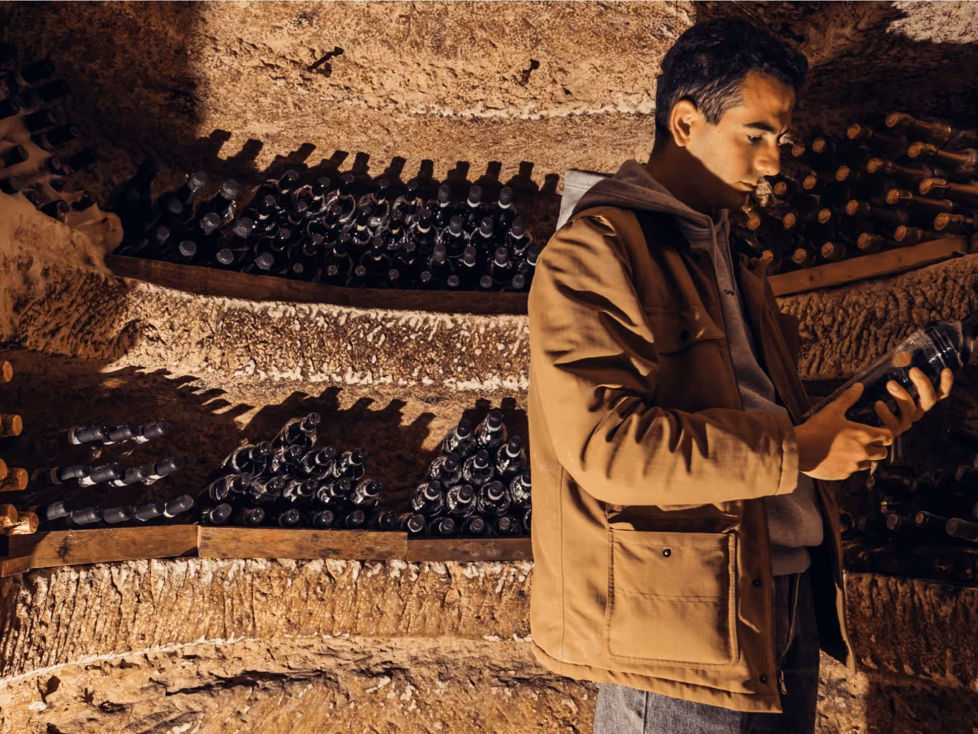 Man examines wine bottle in rustic underground cave near Asti, surrounded by shelves of aged wine bottles.
