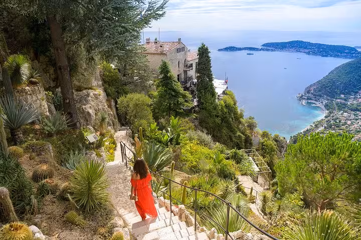 Woman in red dress walking down a lush garden path in Eze Village with stunning coastal views, highlighting a scenic tour stop.