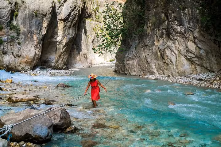 A woman in a red dress carefully crosses a turquoise stream with a rope in scenic Saklıkent Canyon.