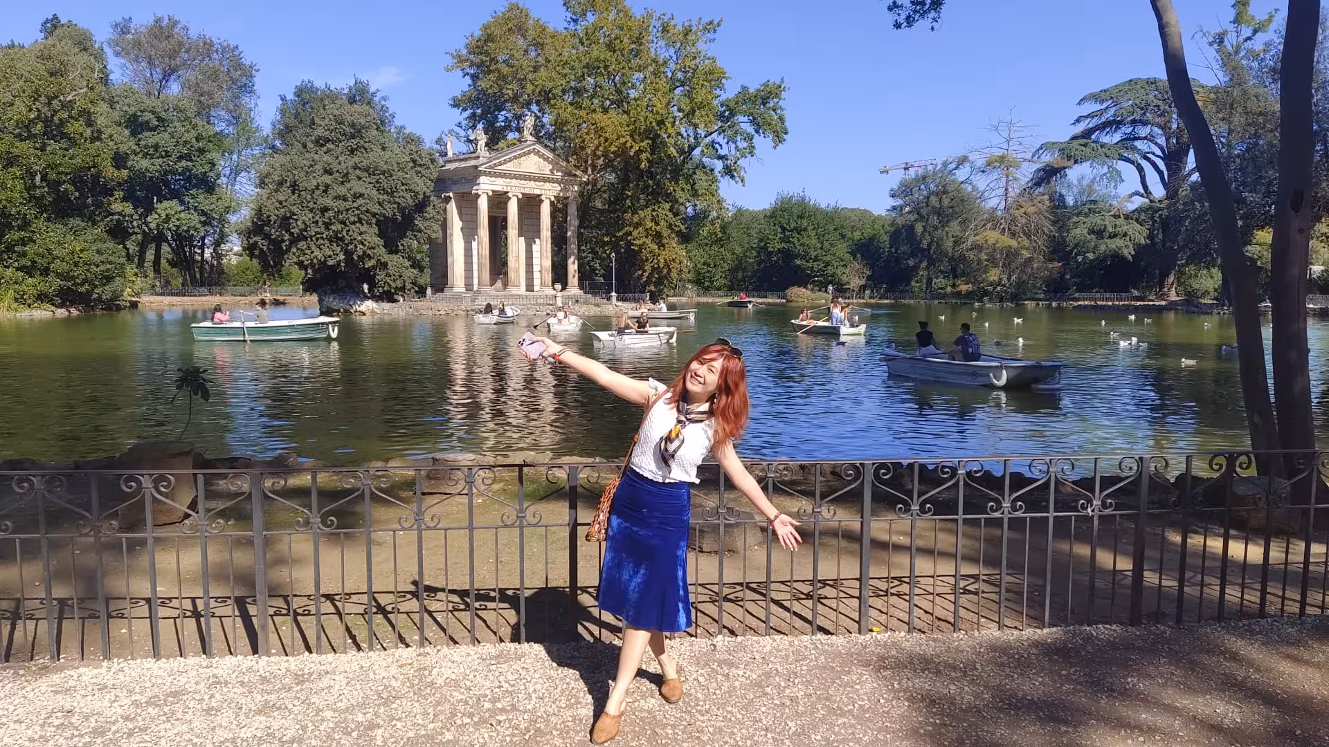 Joyful woman poses by the lake in Villa Borghese, Rome, with Temple of Aesculapius and boating activities in view.