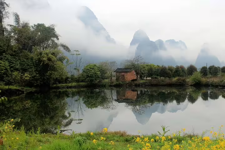 Tranquil Yangshuo landscape with misty karst peaks, a rustic house, and vibrant wildflowers reflected in a serene pond.