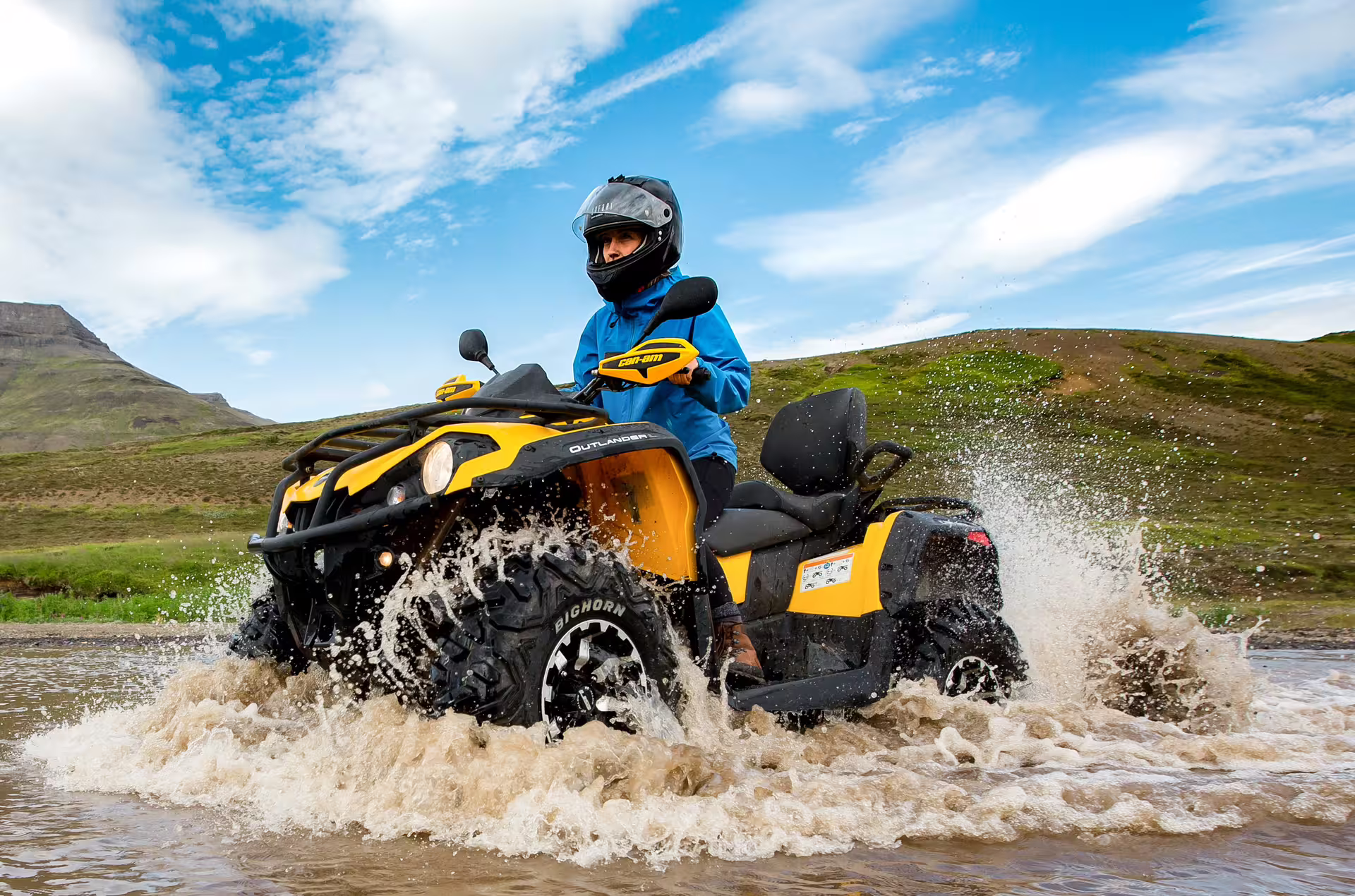 Adventurer rides yellow ATV through water with scenic mountain backdrop on 1 Hour Reykjavik Peak ATV tour in Iceland.